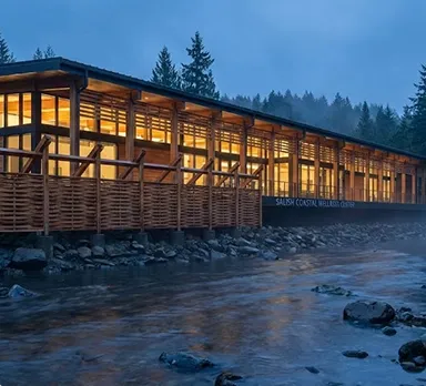 A long, single-storey timber and glass wellness centre lit warmly at dusk, standing on a rocky riverbank below forested hills