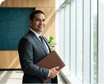A man in a dark suit holds a leather portfolio and smiles, standing in a sunlit corridor lined with floor-to-ceiling windows